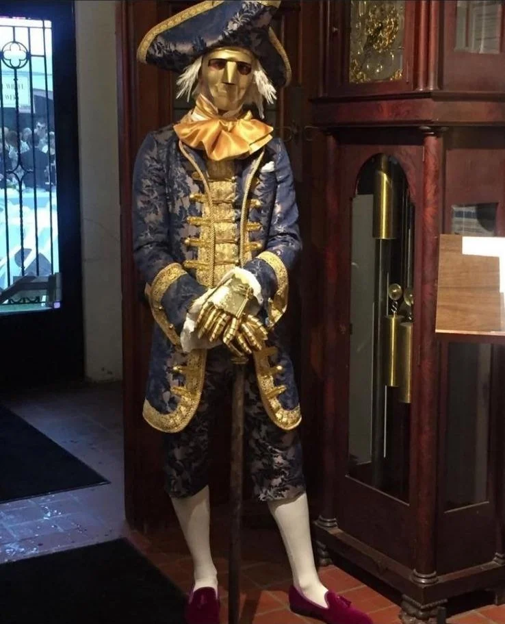 A mannequin dressed in an elaborate costume resembling a royal figure from the 18th century, with a gold mask, blue and gold embroidered coat, lace cuffs, knee-length breeches, white stockings, and red shoes, standing next to a grandfather clock.