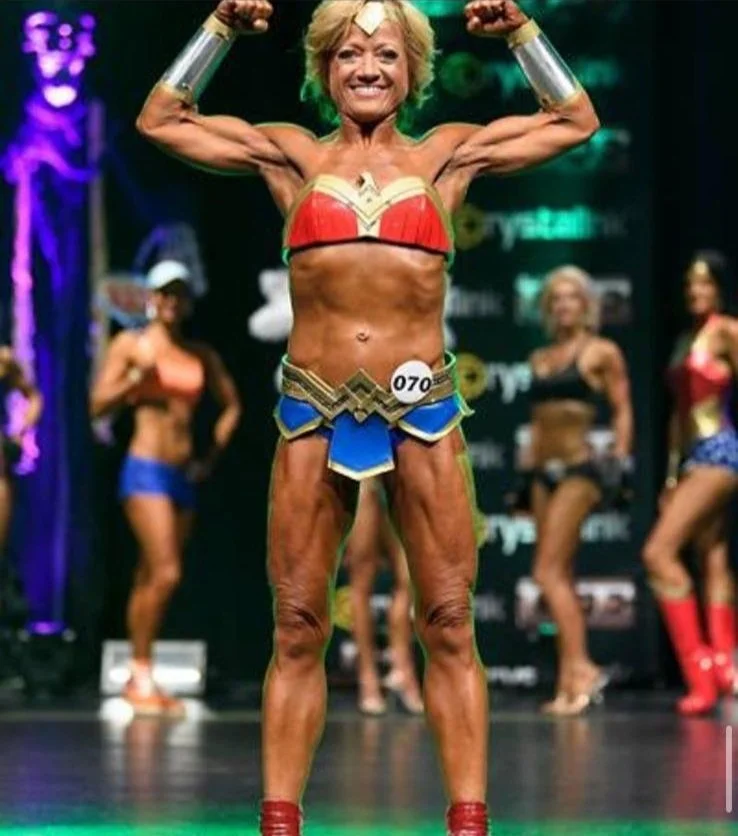 A woman in a Wonder Woman costume flexing her muscles on stage during a fitness competition, with other women in workout outfits in the background.