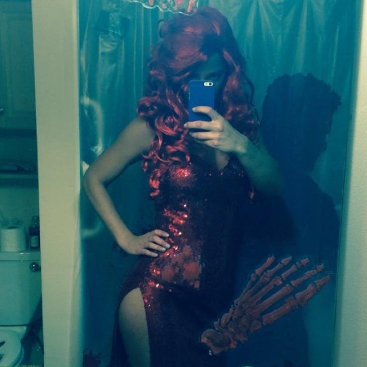 Person with long, curly red hair taking a mirror selfie in a black and red dress with sequins, standing in a bathroom with blue shower curtain. Reflection shows an arm with a skeleton hand tattoo.