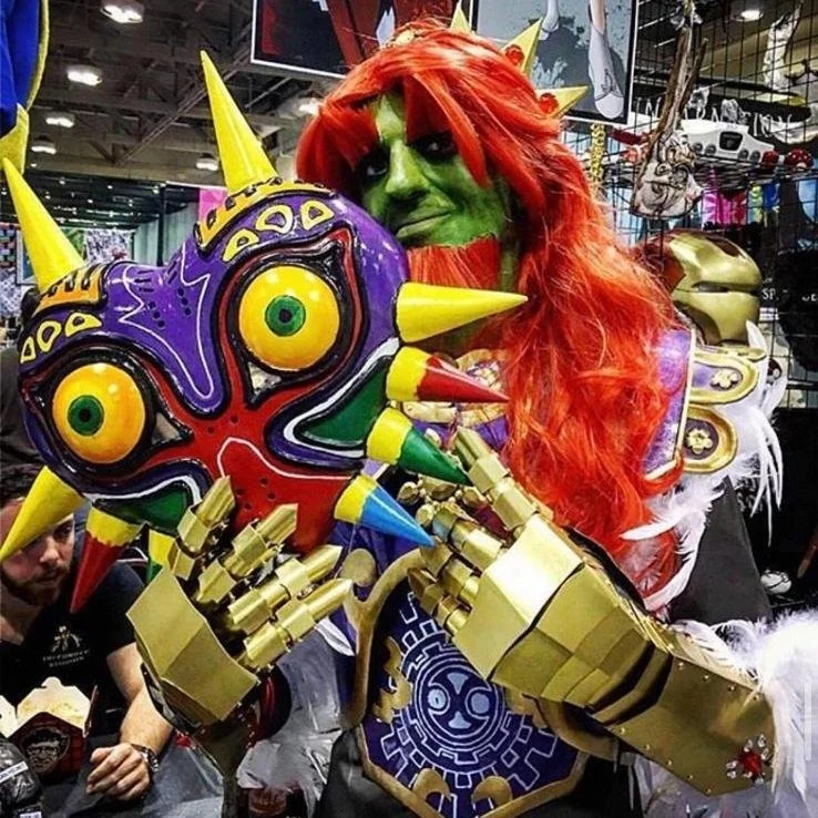 Person dressed as a character with green face paint, red wig, and colorful costume holding a large, ornate mask with spikes in a convention setting.