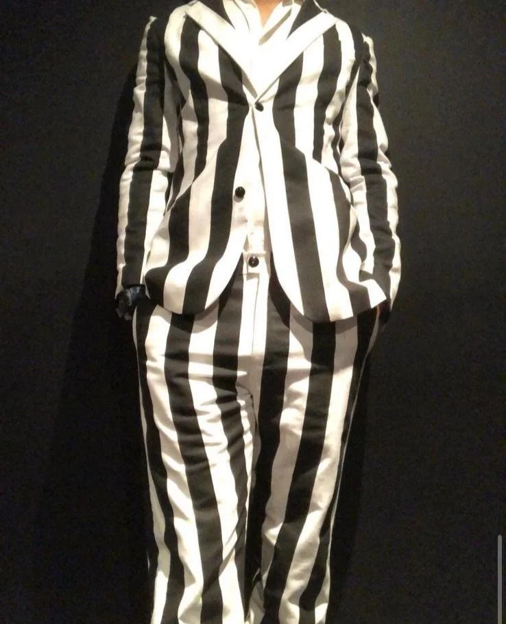 Person wearing a black and white vertically striped suit, including blazer and pants, standing against a dark background