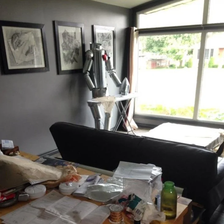 A household room with a large window showing a yard outside. Inside, there is a metallic humanoid robot in the corner near the window, with a small iron board in front of it. The room has a dark couch and a cluttered table with papers, containers, an