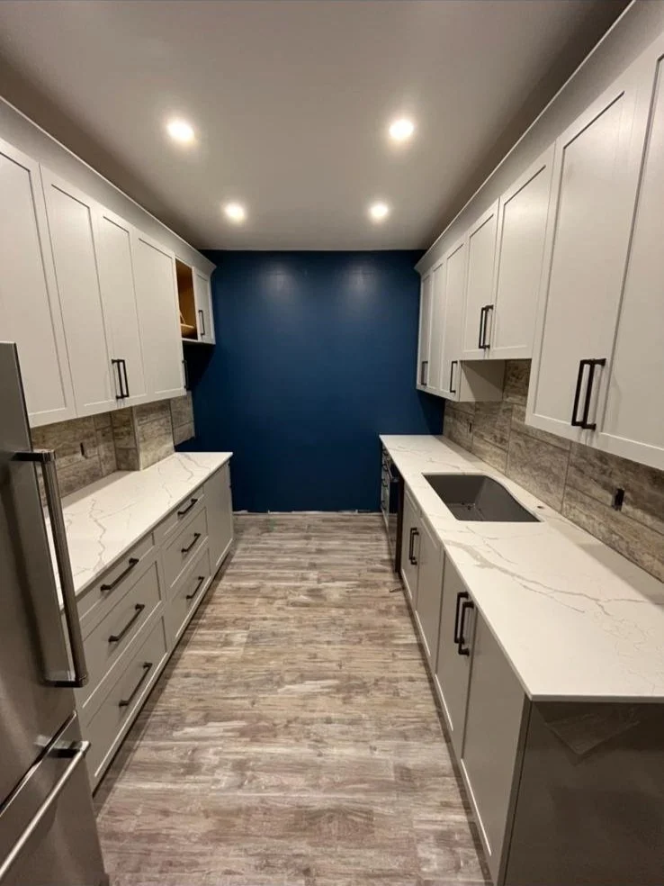 Modern galley kitchen with white cabinets, gray countertops, a blue accent wall at the end, gray wood-like flooring, and recessed ceiling lights.
