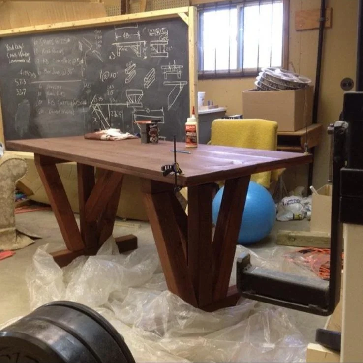 A wooden table in a workshop with black chalkboard in the background, which has sketches and measurements on it. Nearby are chairs, a yellow cushioned chair, a blue exercise ball, and various boxes and items stacked around.