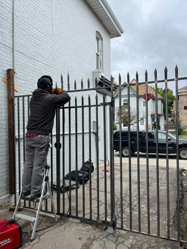 A person standing on a step stool painting a black metal fence in a residential backyard, with a large white house, a black pickup truck, and neighbouring houses visible in the background.
