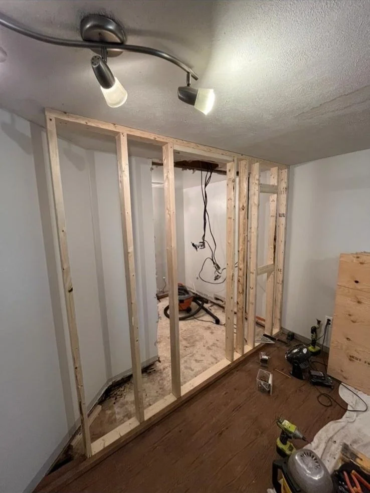 Interior of a room under renovation with exposed wooden framing, construction tools on the floor, and a partial view of a ceiling light fixture.