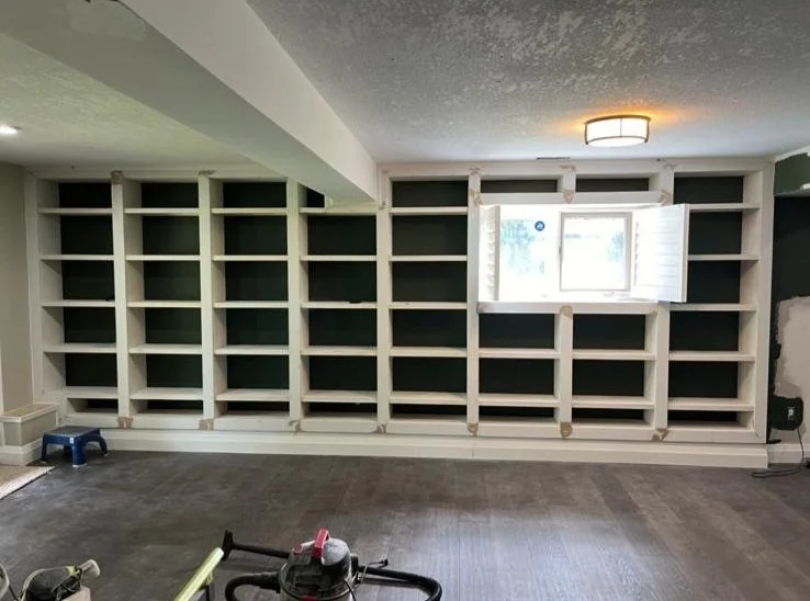 Unfinished built-in bookshelf with white framing and dark backing, featuring a small window with shutters in the middle, in a room with dark flooring and a ceiling light.