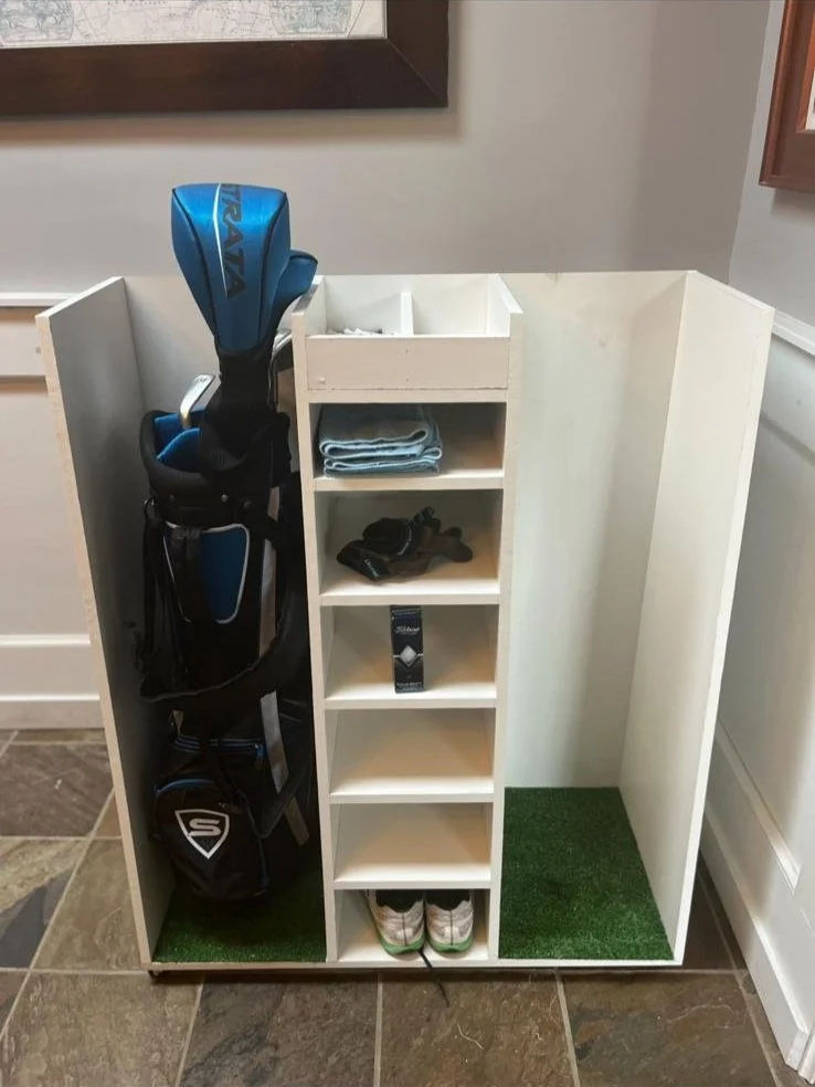 Golf club bag, towels, sunglasses, golf glove, and shoes organized on a white storage unit with artificial grass on the floor.
