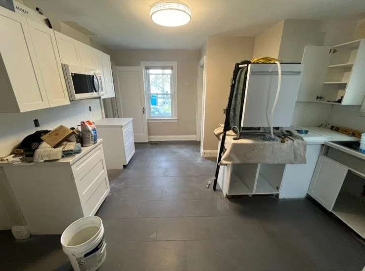 Kitchen under renovation with white cabinets, a microwave, and construction tools and materials on counters and tables.