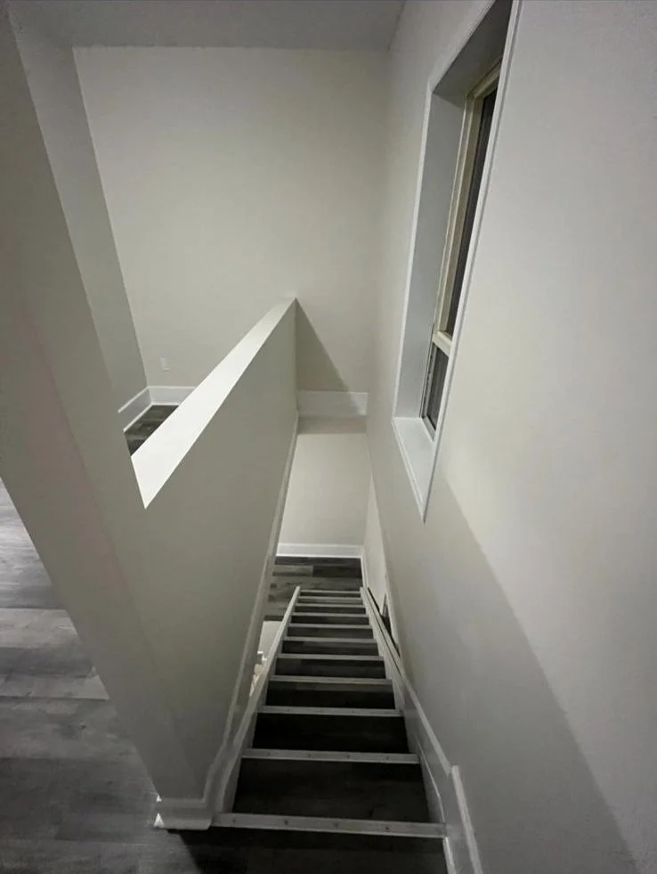 View of an indoor staircase leading down to a lower level, with a half wall on the left side and a window on the right wall. The walls and trim are painted white, and the stairs have dark wood treads with white risers.