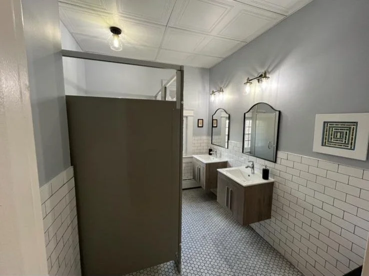 Public bathroom with two sinks, mirrors, and a privacy stall, decorated with white tile wainscoting and light gray walls.