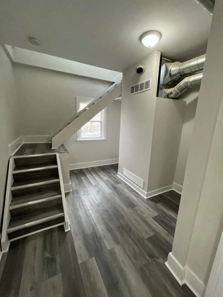 Small room or attic space with sloped ceiling, gray wood flooring, a staircase, a window, and visible HVAC ductwork.
