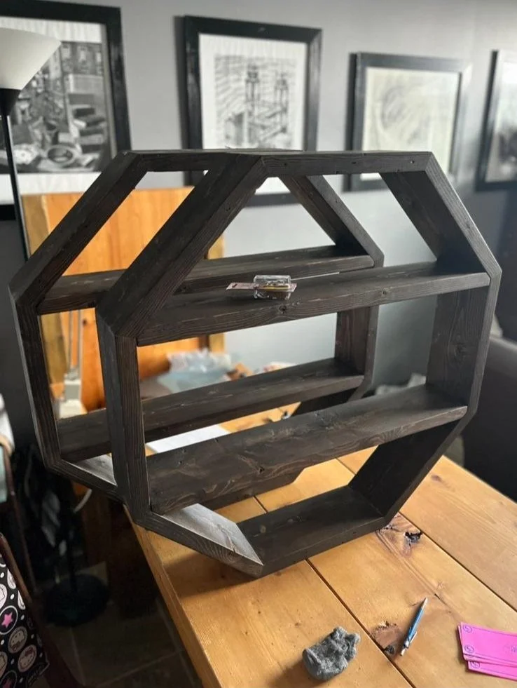 Unfinished black wooden geometric shelf with multiple rectangular and triangular compartments on a wooden table, with tools and materials nearby.