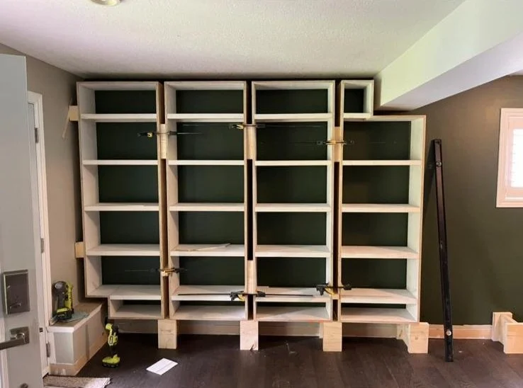 Wooden framing for a built-in bookshelf on an interior wall, with clamps securing sections, in a room with dark hardwood floors and a small window.