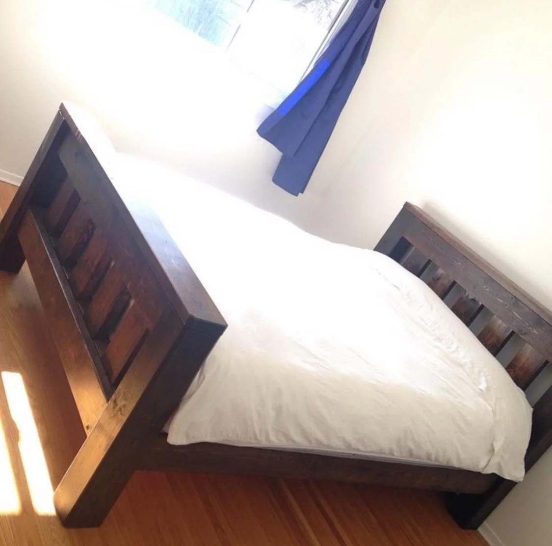 A wooden bed with a white mattress in a sunlit bedroom with white walls, blue curtains, and a window.