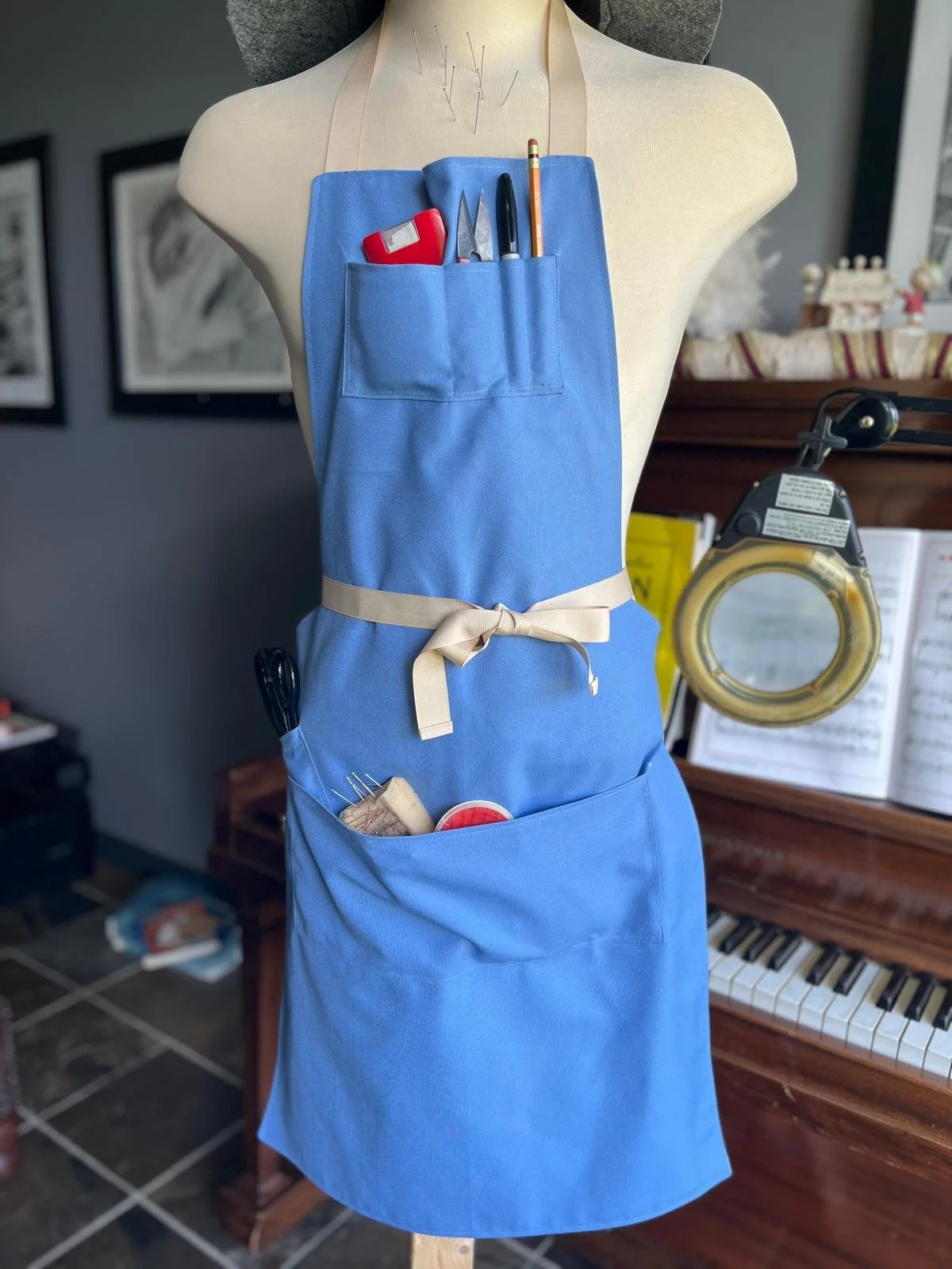 A tailor's dress form wearing a blue apron with various sewing tools inserted into its pockets, including scissors, a seam ripper, a seam gauge, a pencil, and pins.
