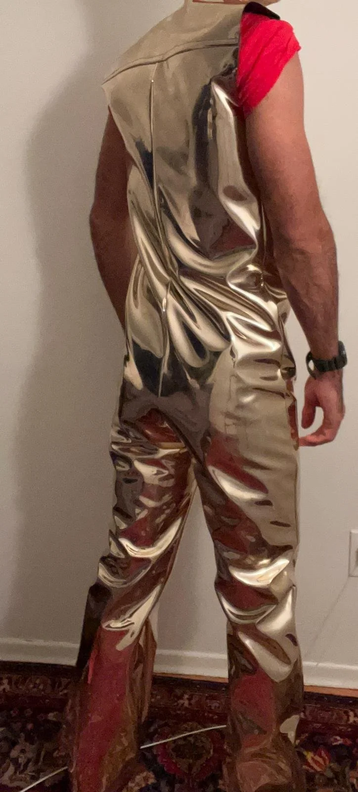 A person wearing a shiny, metallic-looking sleeveless suit, with a red shirt underneath, standing indoors against a plain wall.
