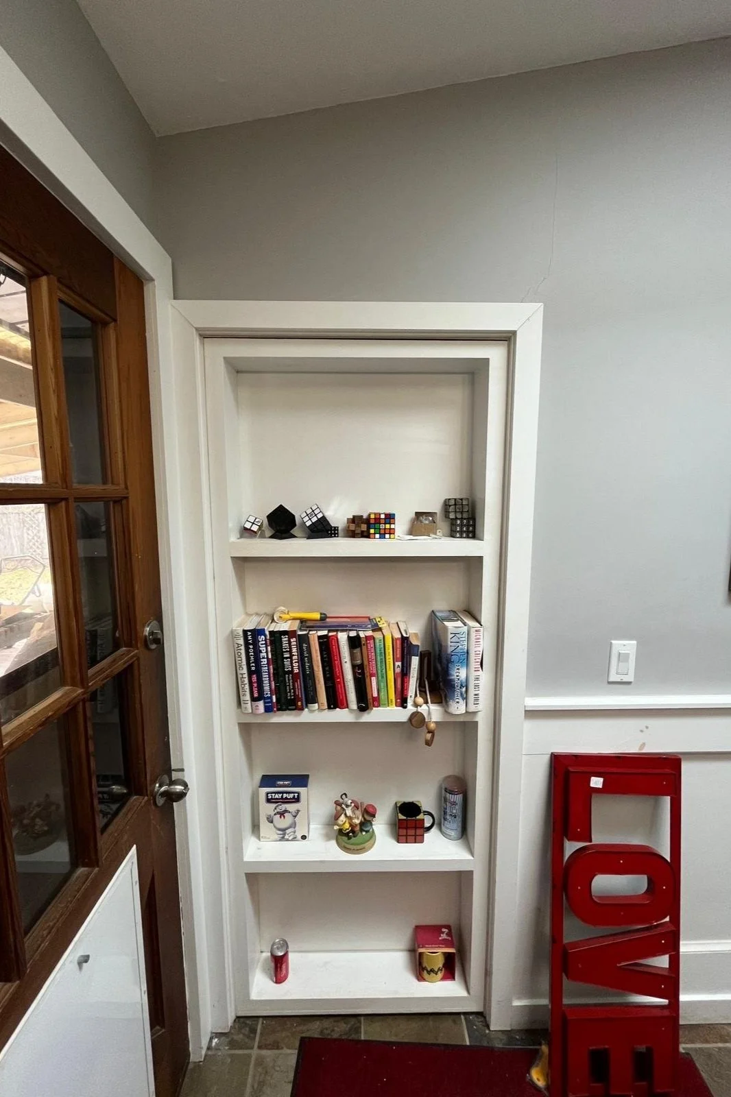 White bookshelf with books, Rubik's cubes, and decorative items in a room with gray walls, a wooden and glass door on the left, and a red 'LOVE' sign on the right side.
