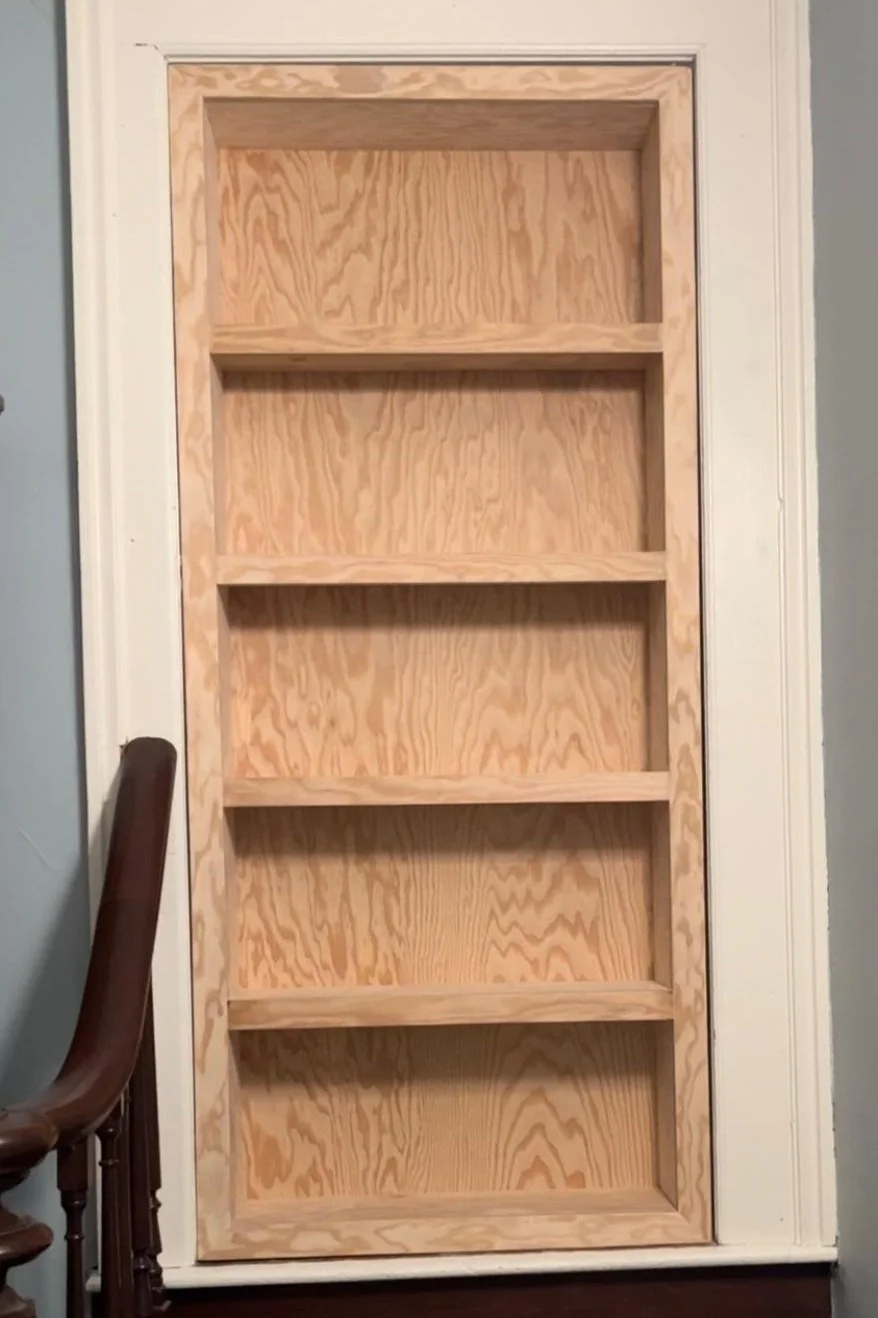 Unfinished secret door wooden bookshelf with five empty shelves, framed in a white door.