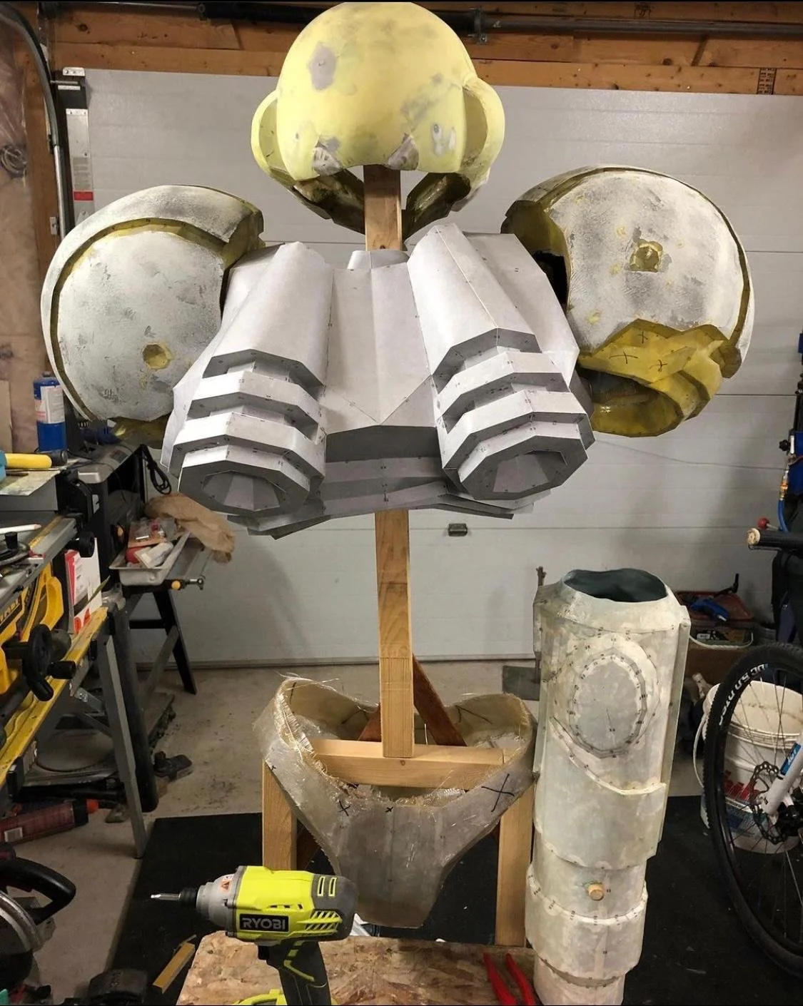 Unfinished robot sculpture in a workshop with a wooden frame, foam, and metal parts. The sculpture has a head, large shoulder parts, and a torso.