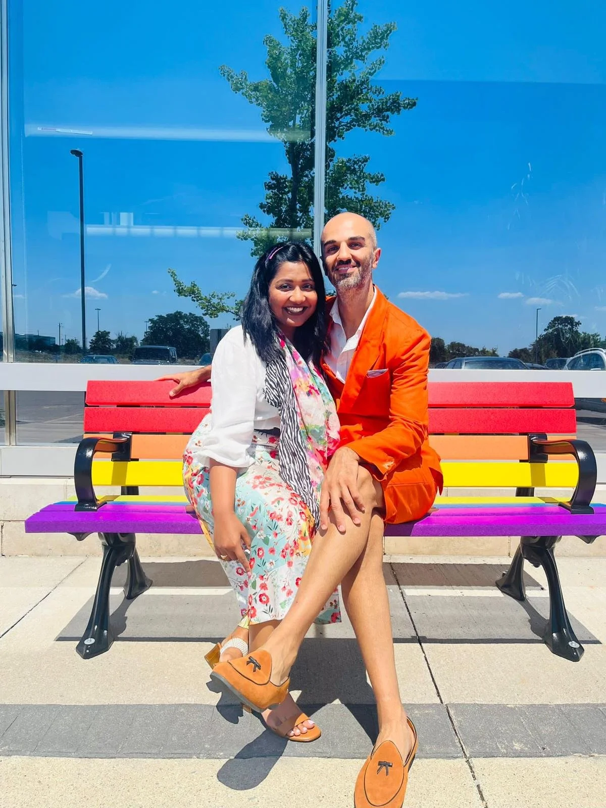 A smiling couple sitting on a colorful park bench outside a building with large glass windows, reflecting a blue sky and trees, on a sunny day.