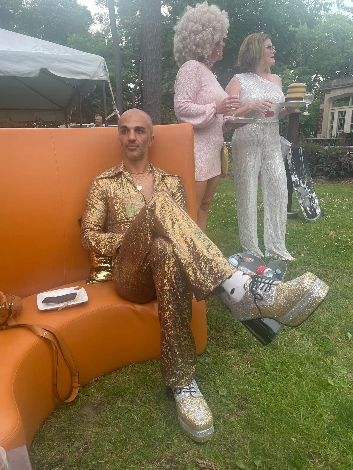 A man dressed in a shiny, gold, and black patterned suit and shoes, sitting on an orange couch at an outdoor gathering, with two women standing behind him, holding plates of food and drinks.