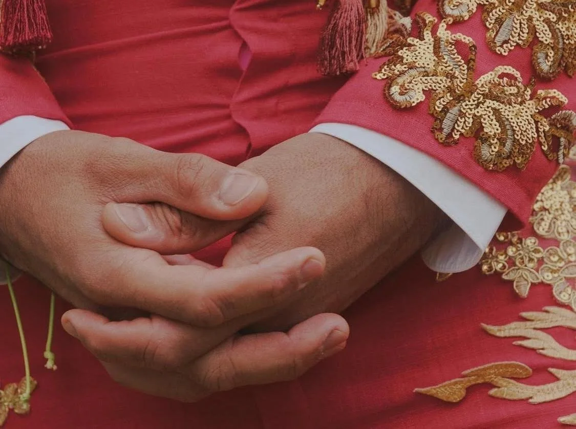 Close-up of two hands clasped, one in a white cuff and the other in a red and gold embroidered fabric, suggesting a handshake or a gesture of unity.