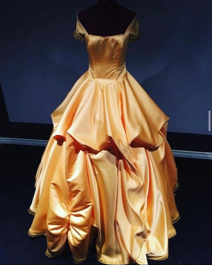 A vintage-style, gold satin ball gown with a fitted bodice and a full, elaborate skirt flounced with pleats and draping.