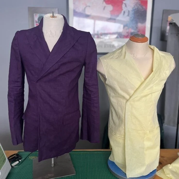 Two dress forms displaying unfinished fashion garments; one in purple fabric with a tailored blazer or coat design, and the other in light yellow fabric with a sleeveless, draped style.