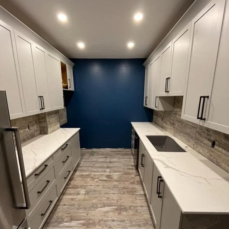 Modern kitchen with white cabinets, marble countertops, gray wood-look flooring, a blue accent wall, and recessed ceiling lights.
