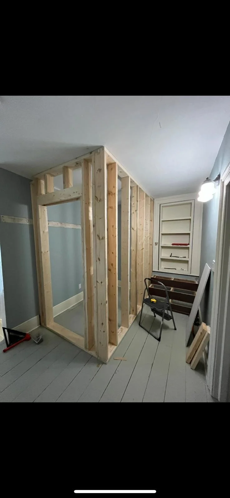 unfinished room with a wooden frame structure, gray painted walls, and built-in shelves in the background, with construction tools on the floor.