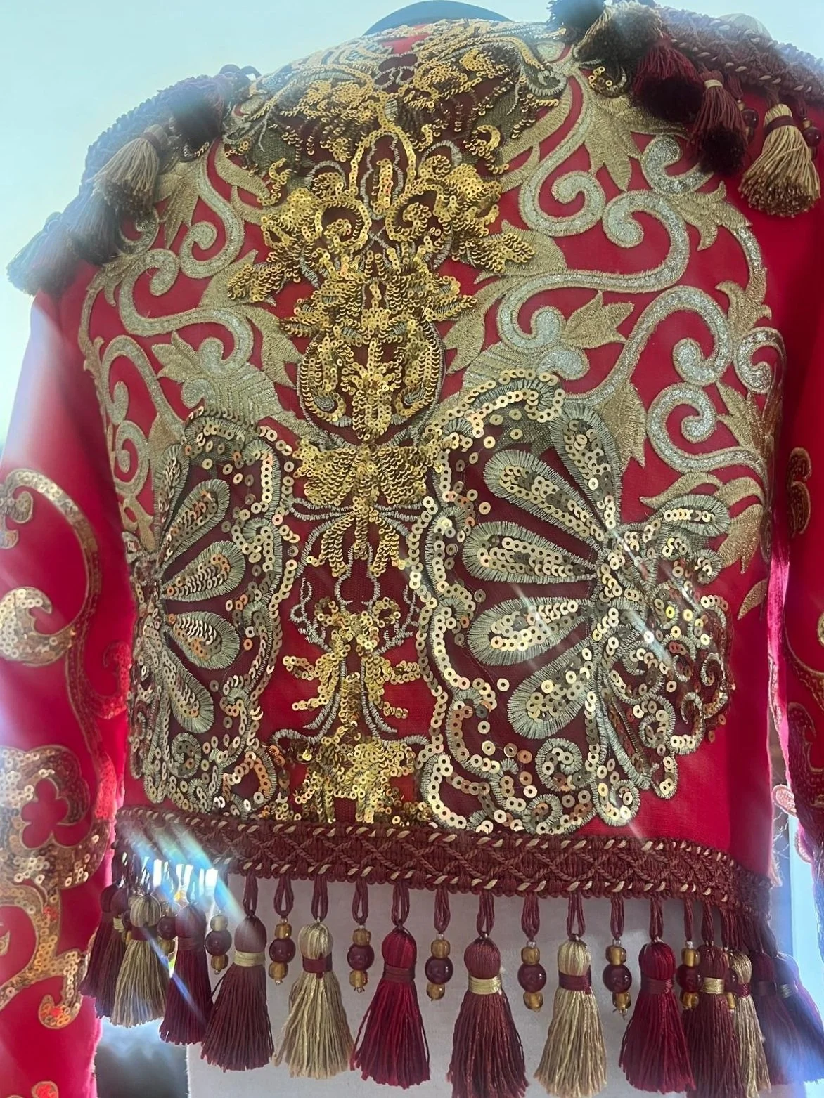Close-up of an ornate, embroidered garment with gold and silver thread on red fabric, decorated with sequins, tassels, and fringes.
