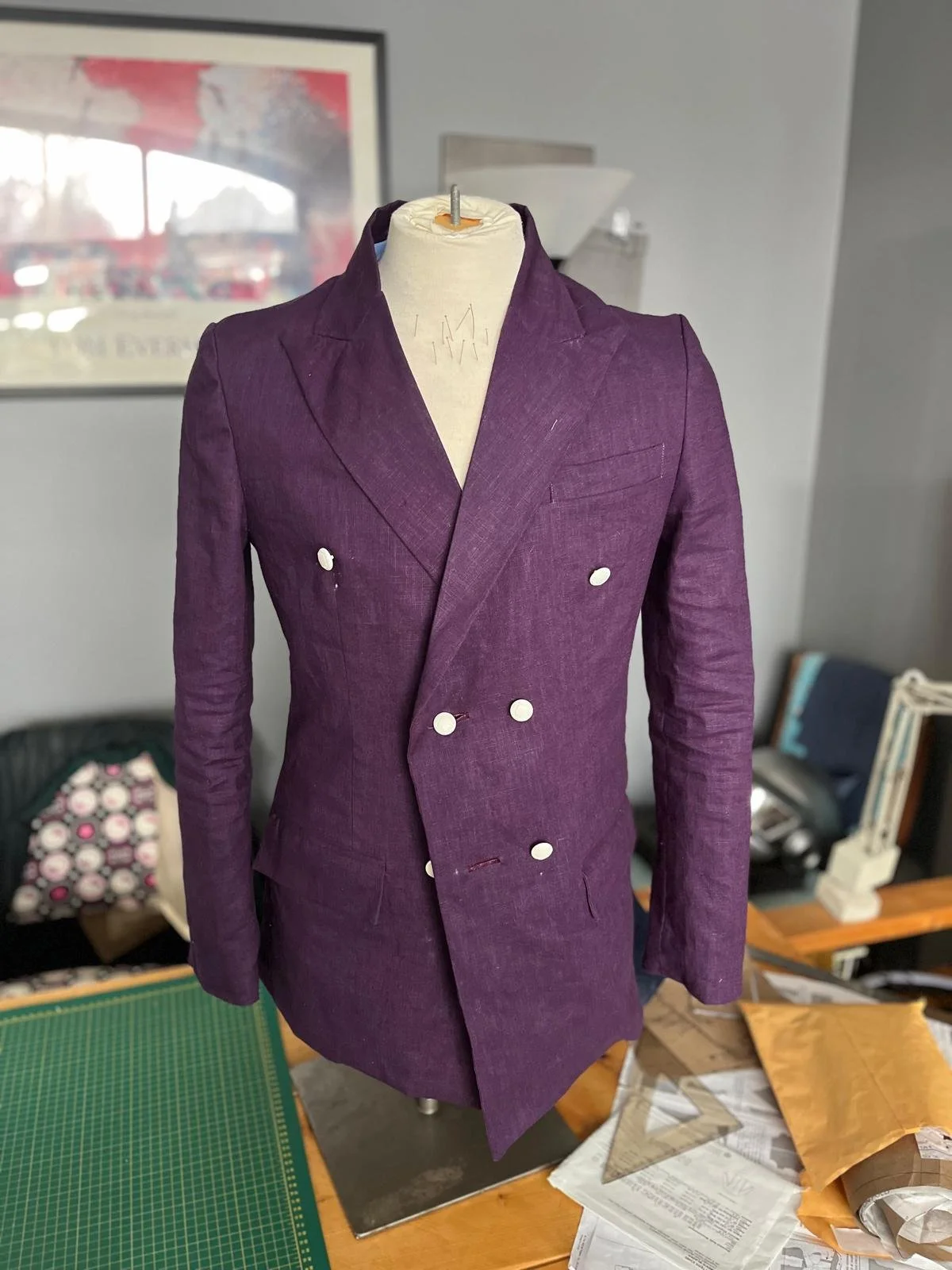 Purple double-breasted blazer with white buttons on a mannequin in a room with framed picture, sewing supplies, and paperwork.