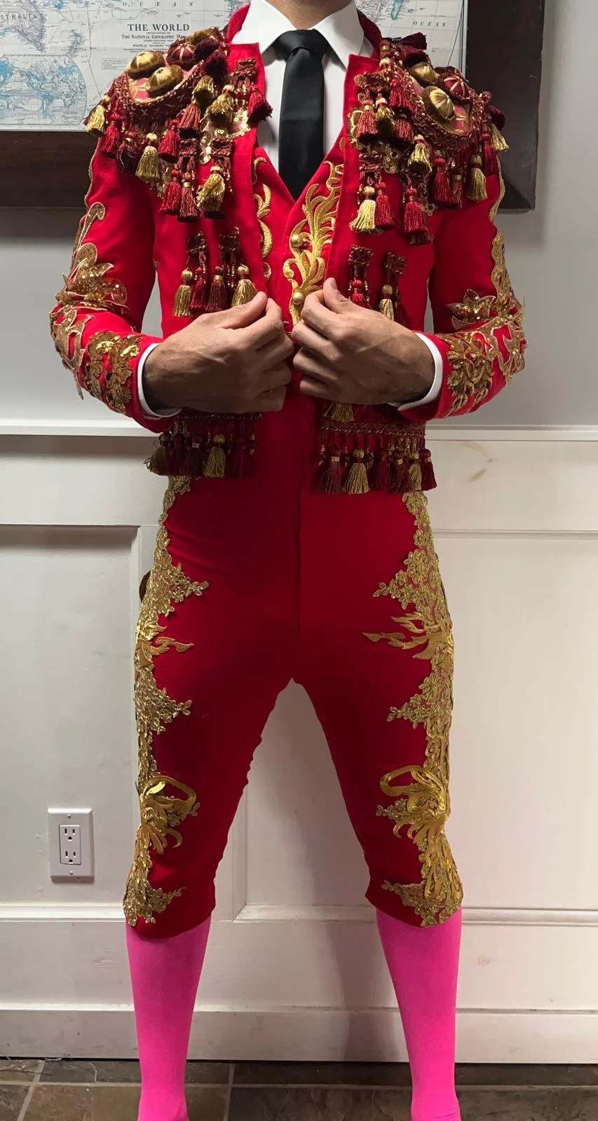 A person dressed in a red and gold ornate matador-style costume with intricate embroidery and tassels, wearing a white shirt, black tie, and pink socks, standing indoors with a world map in the background.