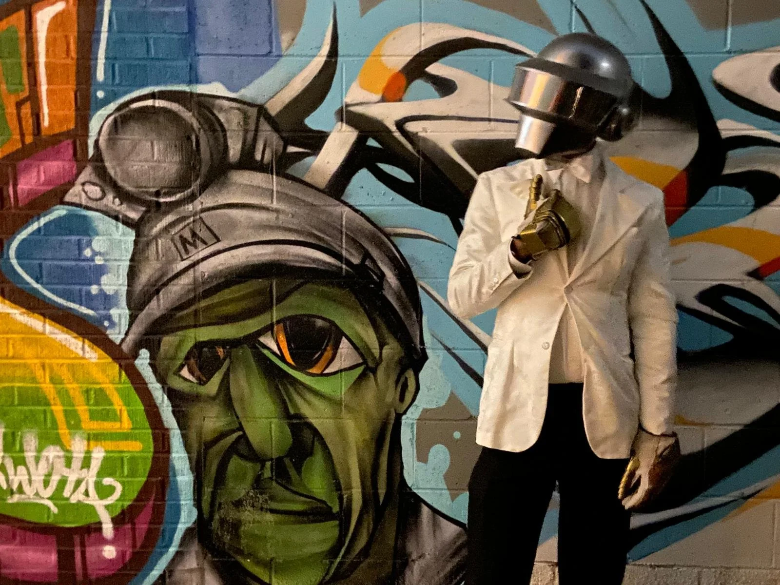 Person dressed in a white blazer and black pants, wearing a metallic mask, standing in front of colorful graffiti art on a brick wall. The graffiti features a green-faced character with a riot helmet and an eye that appears to be looking to the side.