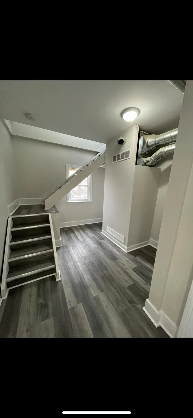 Interior of a modern room with gray wood-like flooring, white walls, a small staircase with three steps, a window letting in natural light, and exposed ductwork near the ceiling.
