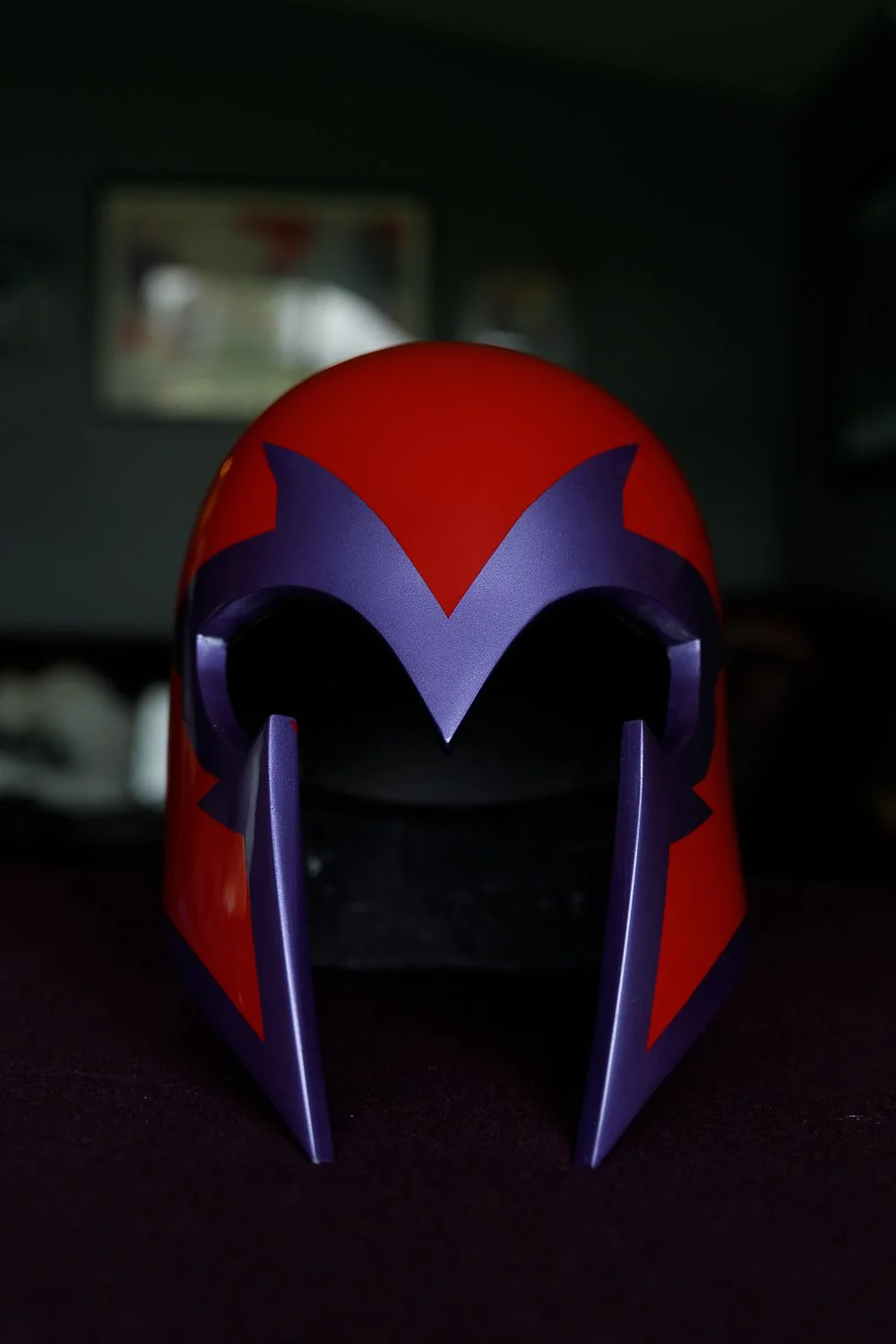 A red and purple superhero mask with a large purple 'V' shape over the eye holes, sitting on a dark surface in a dimly lit room.