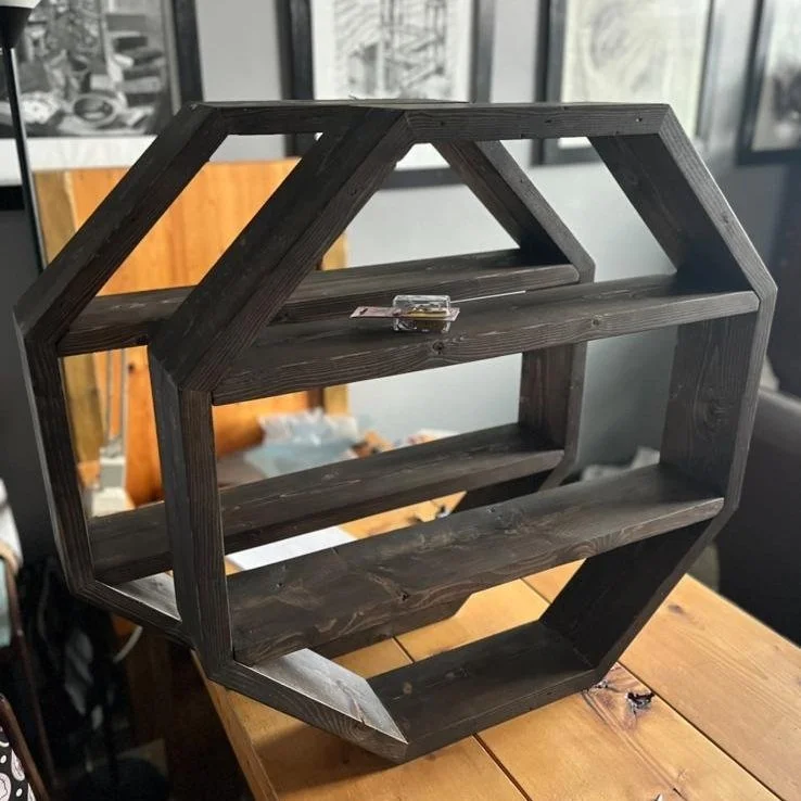 Unfinished dark wood hexagonal bookshelf with three shelves on a wooden table.