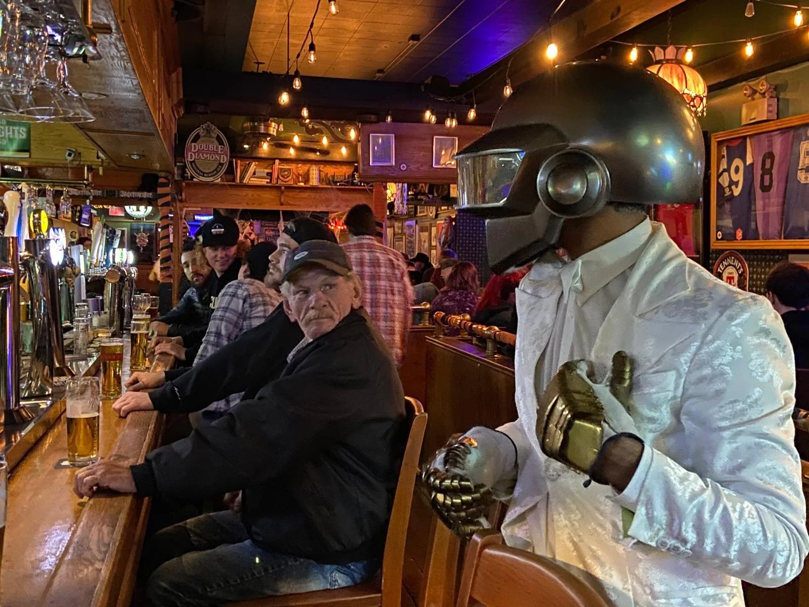 A person in a white shirt with a shiny black helmet and gold gloves stands at a bar in a lively pub, surrounded by patrons sitting and socializing.