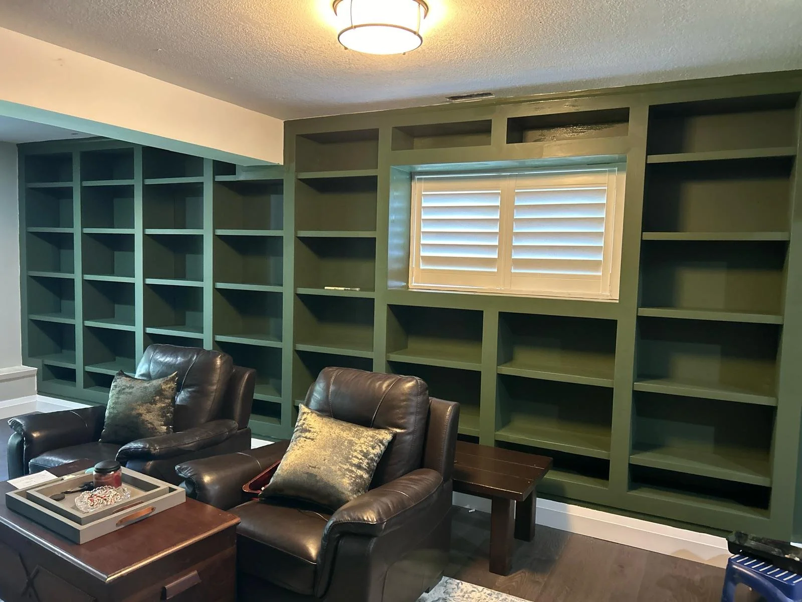 Living room with dark brown leather chairs, a window with white shutters, and a large built-in green bookshelf that is mostly empty.