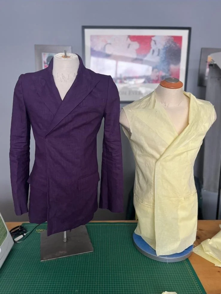 Two dress mannequins, one dressed in a purple jacket and the other in an unfinished yellow garment, on a green cutting mat in a sewing workspace.