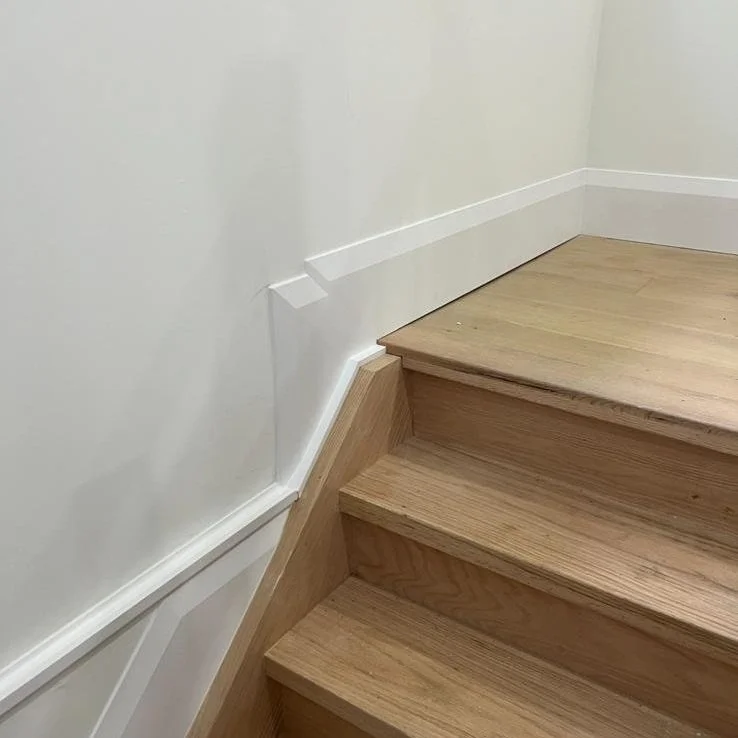 Close-up of a staircase with wooden steps and white painted walls, featuring a corner trim and a running baseboard along the wall.