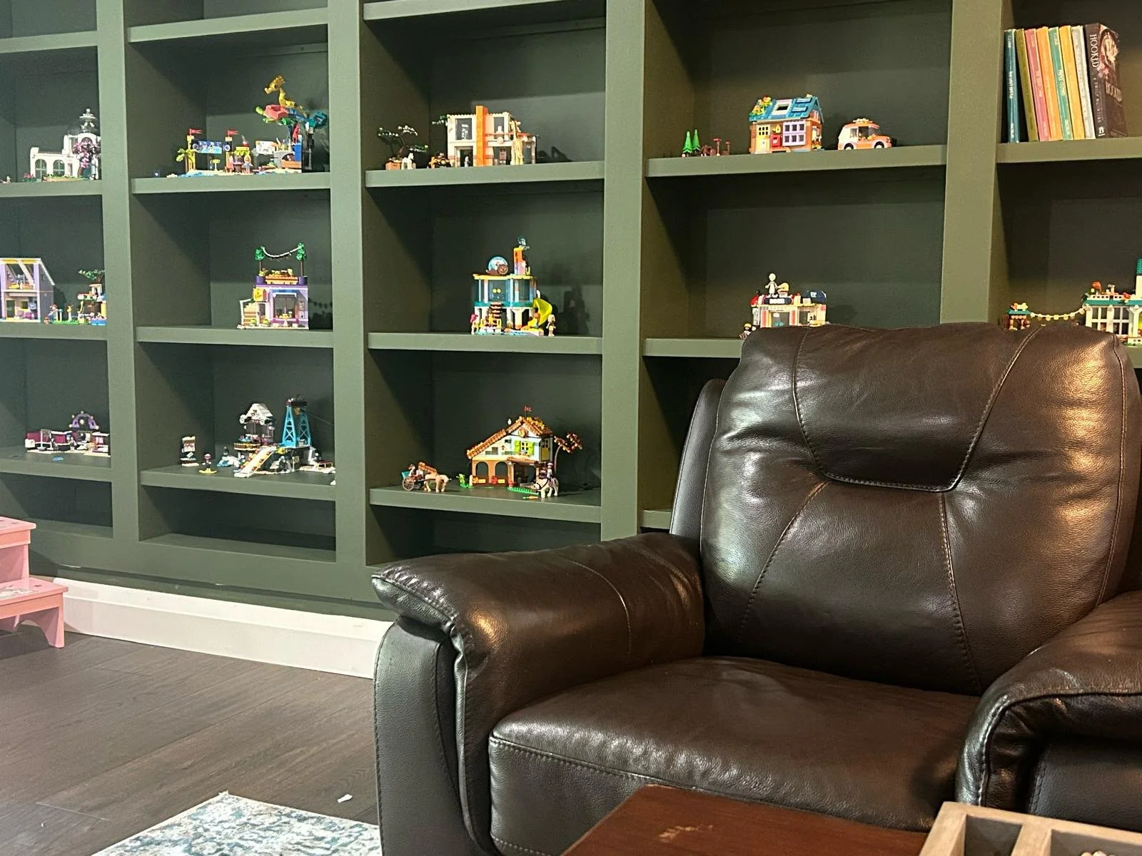 Green bookshelf filled with LEGO sets, next to a brown leather armchair.