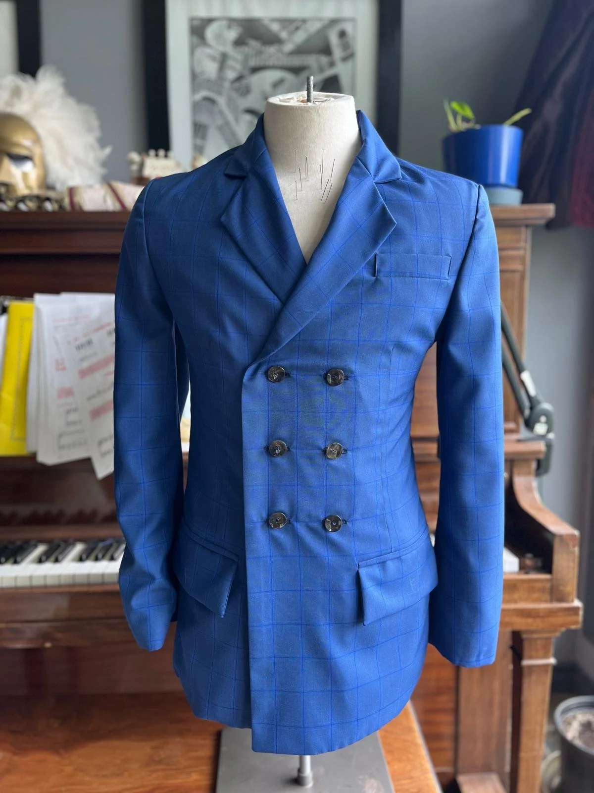 A mannequin dressed in a double-breasted blue plaid suit jacket inside a room with a wooden piano and various decorative items.