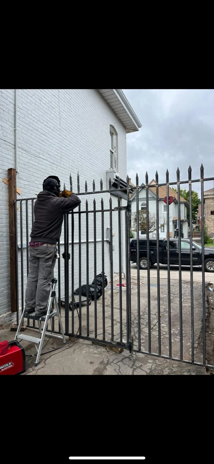 A person working on a black metal gate attached to a white brick building, using a welder while standing on a step ladder, with a black truck parked on the street in the background.