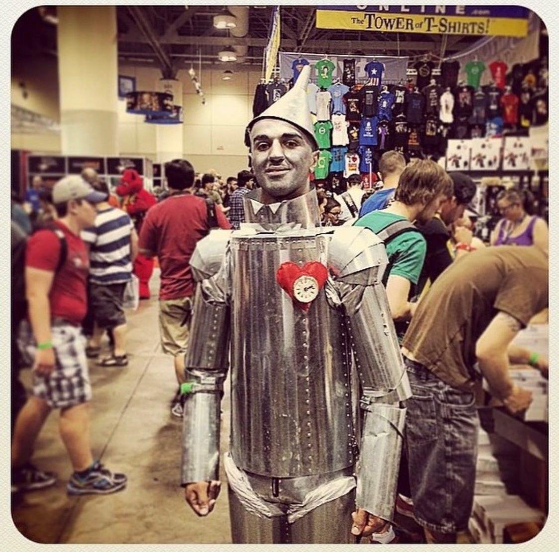 A person dressed as the Tin Man from The Wizard of Oz, with a metallic suit and a cone-shaped hat, standing in a crowded convention or store. The person has makeup to resemble a face and is wearing a heart-shaped clock on chest.