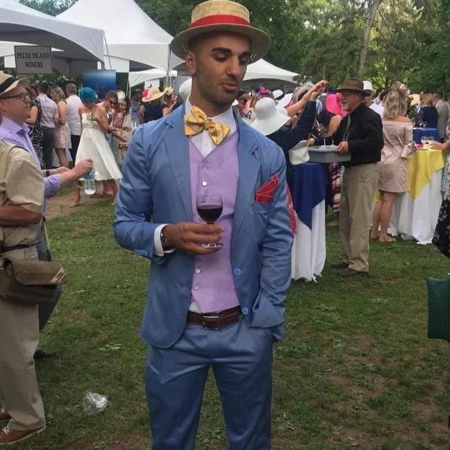 Man in a pastel blue suit and straw hat holding a glass of red wine at an outdoor event with tents and people in the background.