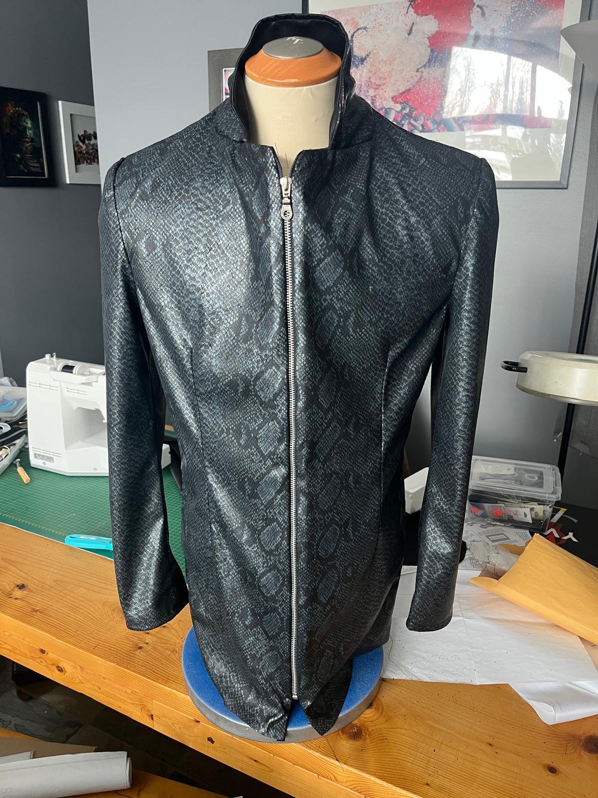Black faux leather jacket with a snakeskin pattern on a mannequin, displayed on a wooden table in a craft room.