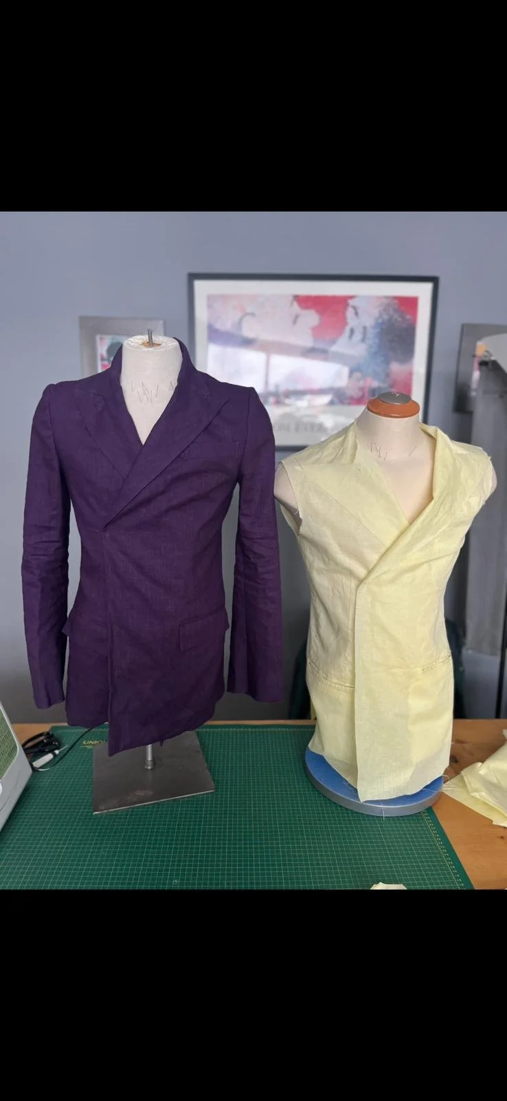 Two dress forms on a work table, one with a purple tailored jacket and the other with a pale yellow sleeveless shirt under construction, in a design studio with wall art in the background.
