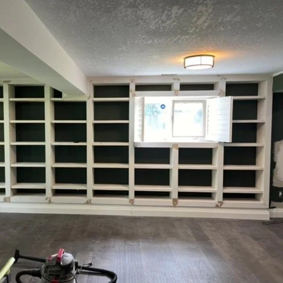 Room with a large built-in open shelving unit and a small window with shutters, under a ceiling light fixture, with a vacuum cleaner partially visible on the floor.